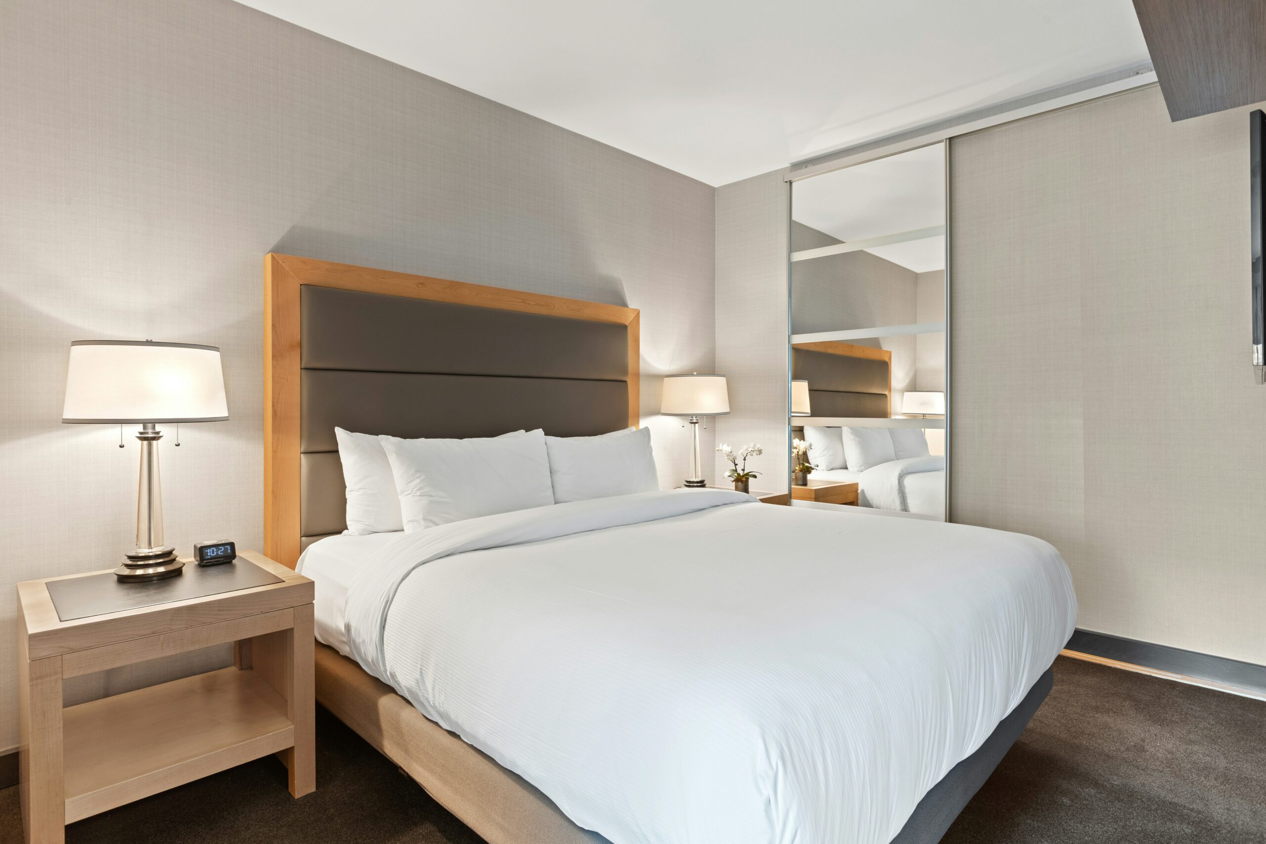 A modern hotel bedroom featuring a large bed with white linens, a wooden headboard, a bedside lamp, and a mirror on the wall. The interior design is contemporary with neutral colors.